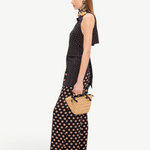 A person in profile wears a black Loch Sleeveless Crepe Button Up with matching wide-leg trousers featuring a tan polka dot pattern, creating an elegant look while carrying a small woven straw handbag.