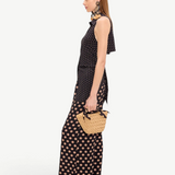 A person in profile wears a black Loch Sleeveless Crepe Button Up with matching wide-leg trousers featuring a tan polka dot pattern, creating an elegant look while carrying a small woven straw handbag.