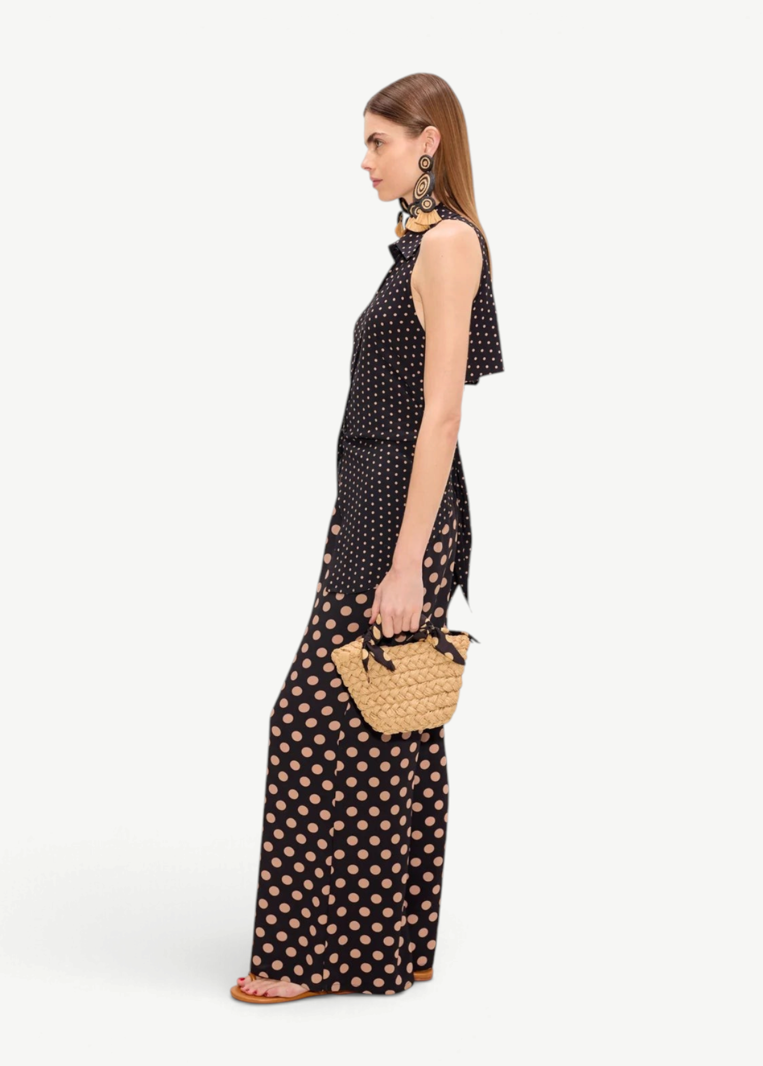 A person in profile wears a black Loch Sleeveless Crepe Button Up with matching wide-leg trousers featuring a tan polka dot pattern, creating an elegant look while carrying a small woven straw handbag.