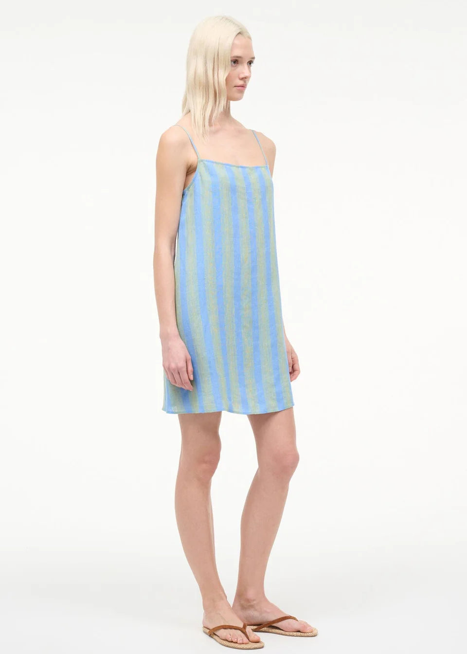 A person wearing a blue and light green striped Laura Mini Dress with spaghetti straps and brown flip-flops, perfect for warm-weather dressing.