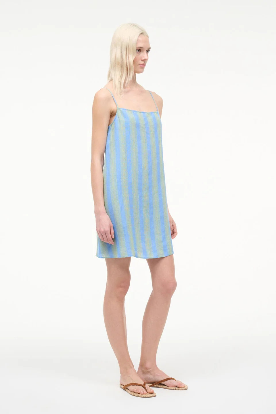 A person wearing a blue and light green striped Laura Mini Dress with spaghetti straps and brown flip-flops, perfect for warm-weather dressing.