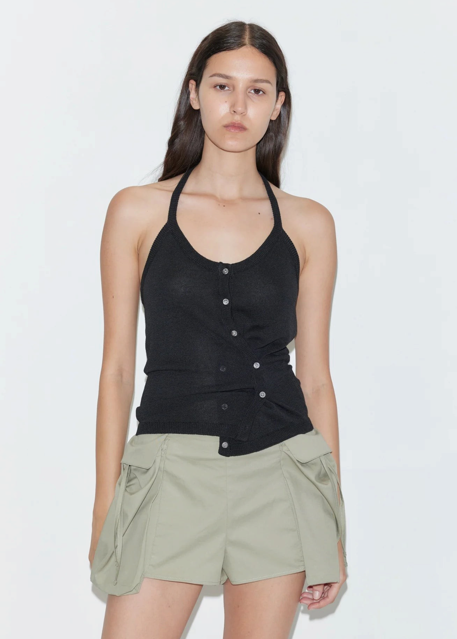 A person wears a Cross Top featuring a fitted knit halter design with an asymmetrical button-front closure and adjustable tie straps, paired with olive green shorts.