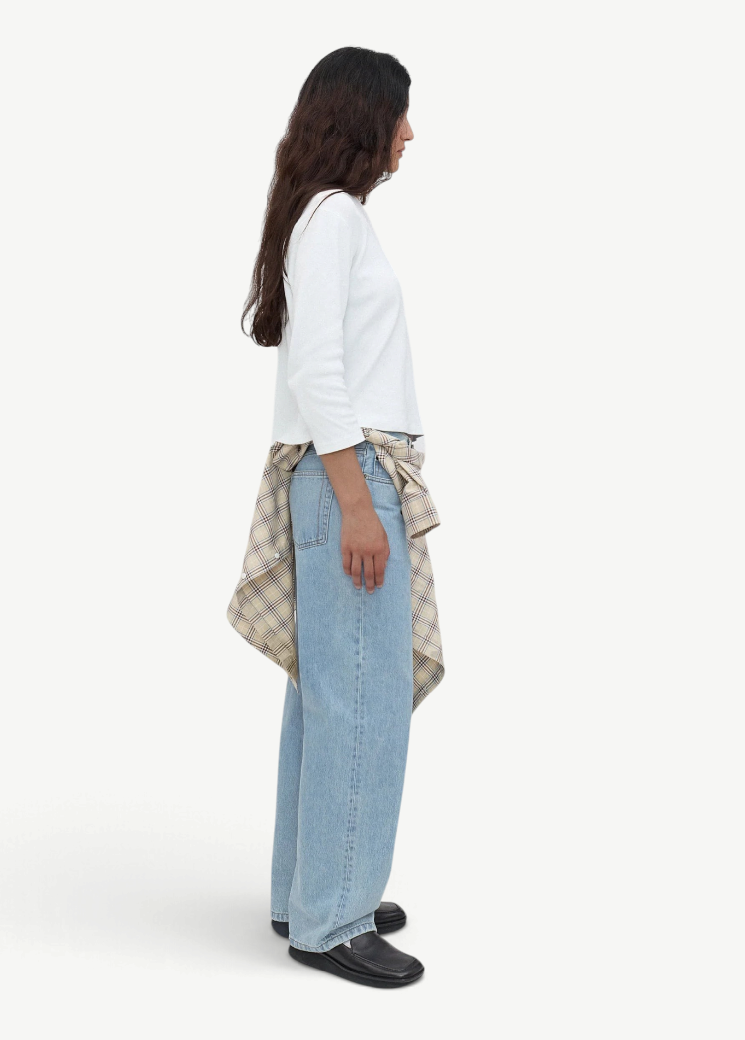 A person stands sideways wearing a white long-sleeve top, black shoes, and light blue Everyday Jeans with a relaxed silhouette. A plaid shirt is tied around their waist, and the background is plain and light gray.
