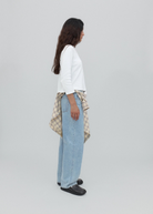 A person stands sideways wearing a white long-sleeve top, black shoes, and light blue Everyday Jeans with a relaxed silhouette. A plaid shirt is tied around their waist, and the background is plain and light gray.