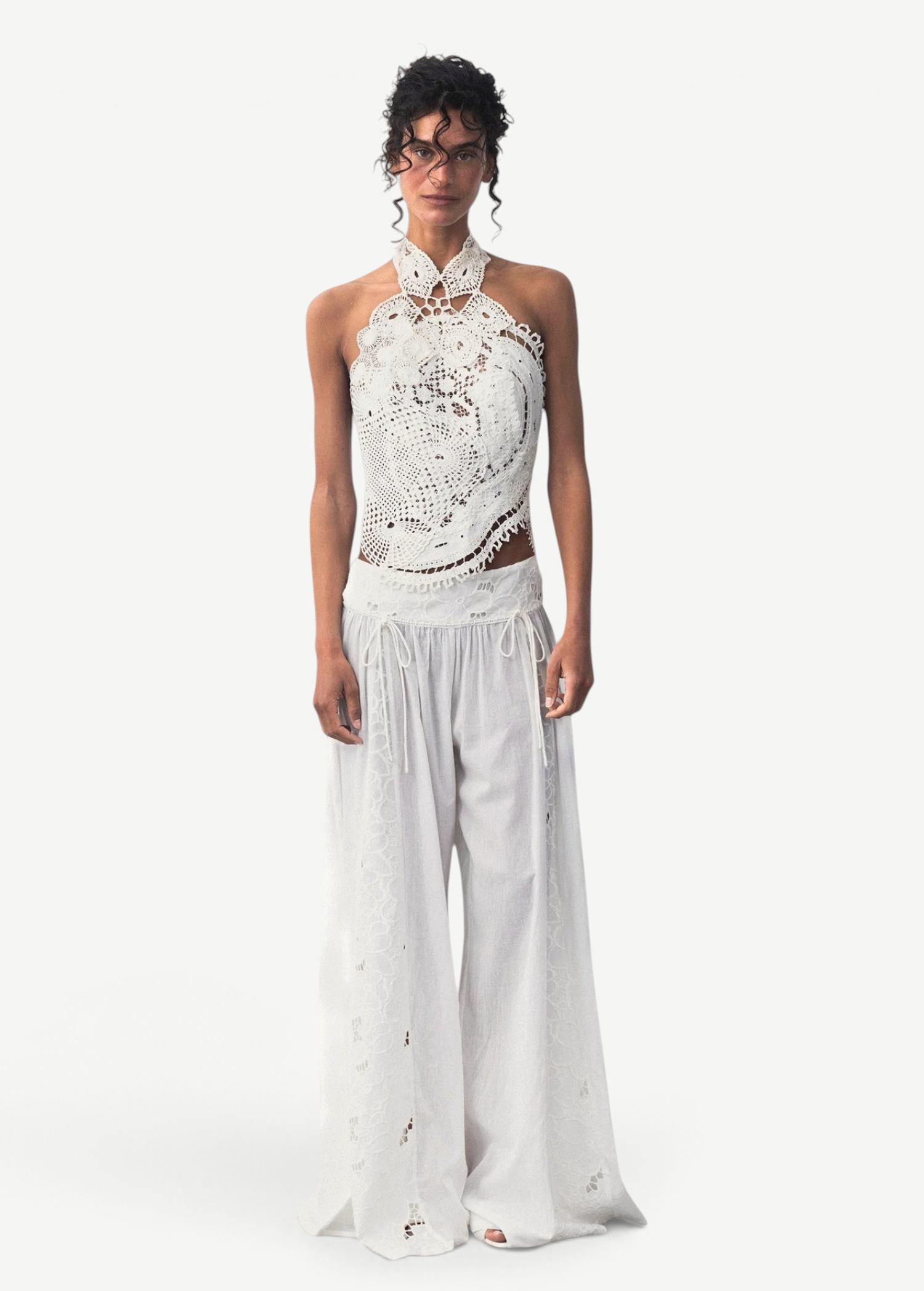 Model wearing KABE SHOP INC Menorca white cotton trousers with intricate lace detailing and a matching lace halter top.