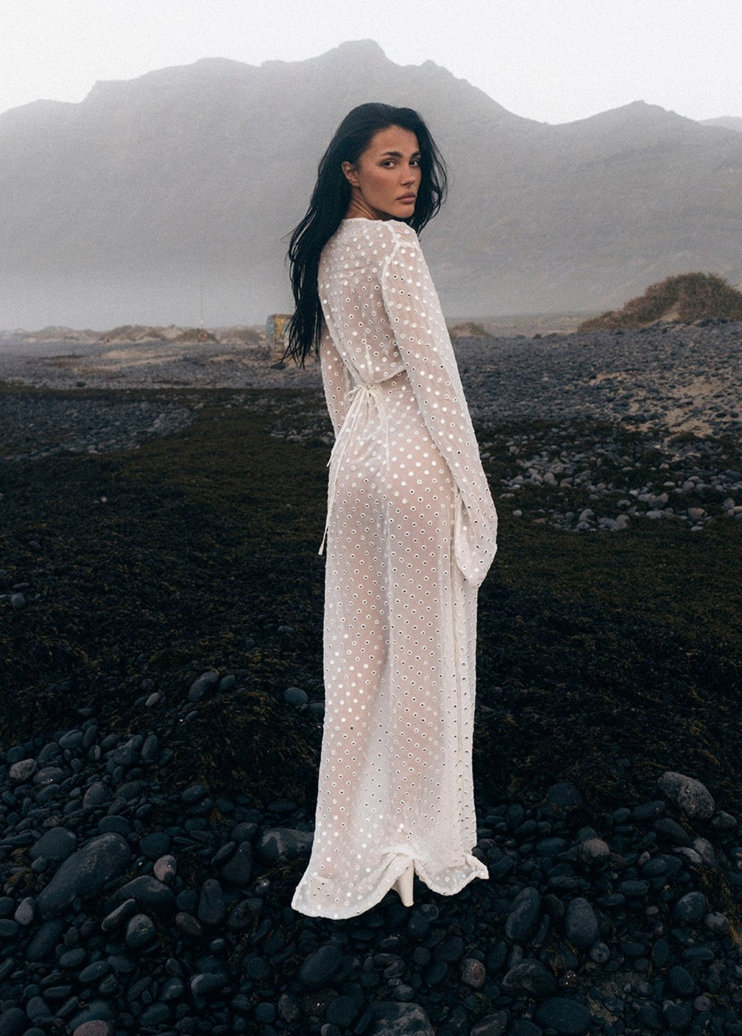 Model wearing a KABE SHOP INC Mirror Dress in sheer white fabric with silver embellishments on a rocky beach