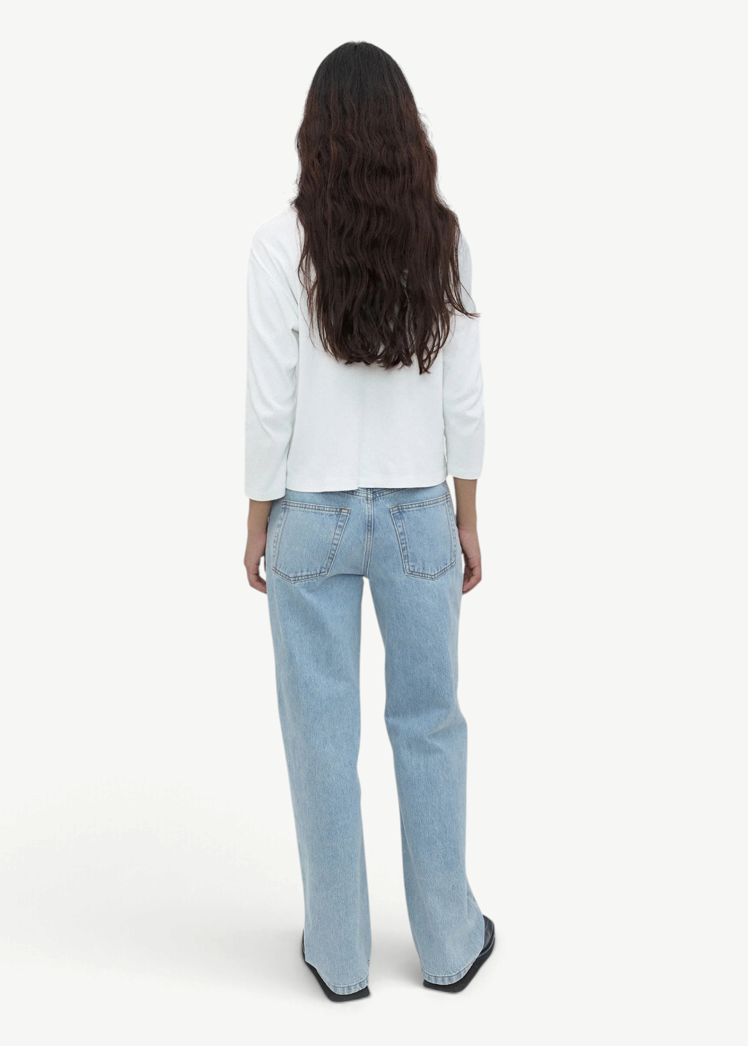 A person with long, wavy dark hair is standing facing away, wearing a white long-sleeve top, versatile light blue Everyday Jeans, and black shoes against a plain light gray background.