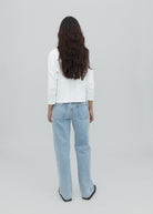 A person with long, wavy dark hair is standing facing away, wearing a white long-sleeve top, versatile light blue Everyday Jeans, and black shoes against a plain light gray background.