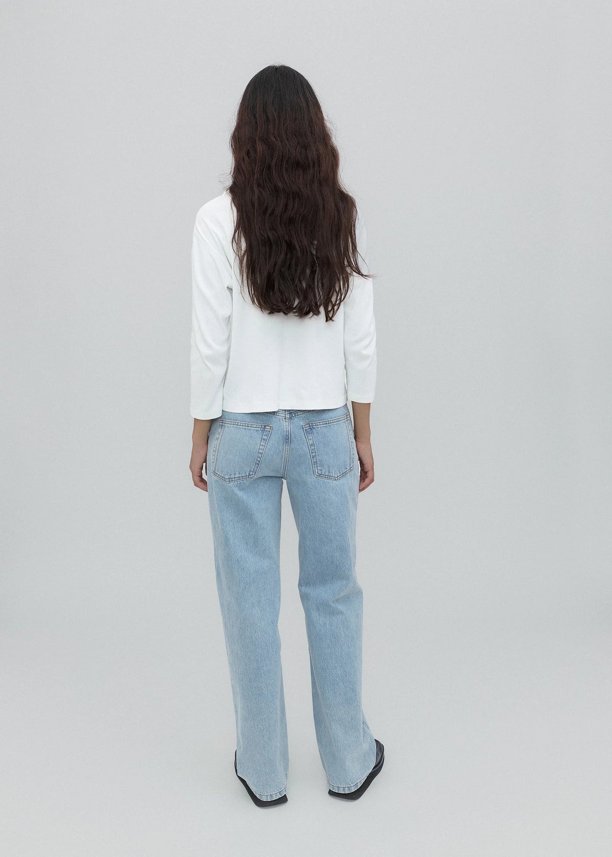 A person with long, wavy dark hair is standing facing away, wearing a white long-sleeve top, versatile light blue Everyday Jeans, and black shoes against a plain light gray background.