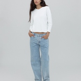 A person with long dark hair stands against a plain light gray background, wearing a white long-sleeve shirt, black shoes, and the Everyday Jeans in light blue—a loose fit perfect for everyday wear—with hands in their pockets.
