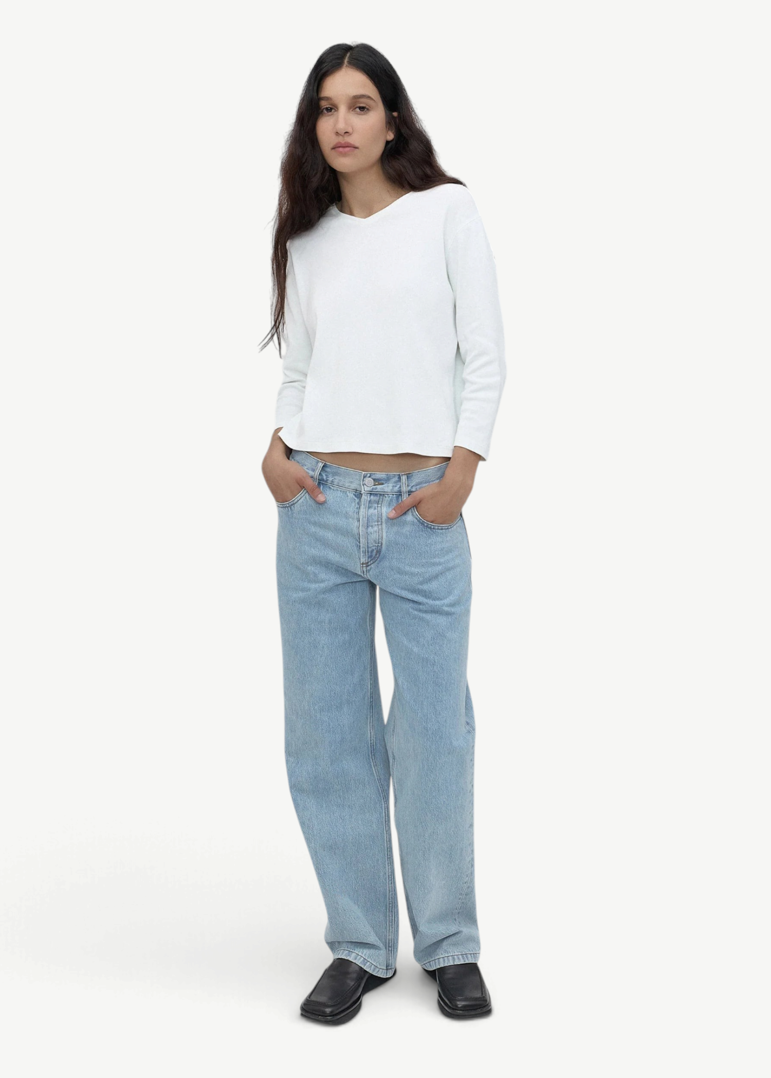 A person with long dark hair stands against a plain light gray background, wearing a white long-sleeve shirt, black shoes, and the Everyday Jeans in light blue—a loose fit perfect for everyday wear—with hands in their pockets.
