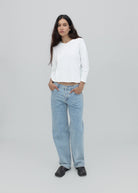 A person with long dark hair stands against a plain light gray background, wearing a white long-sleeve shirt, black shoes, and the Everyday Jeans in light blue—a loose fit perfect for everyday wear—with hands in their pockets.