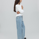 A woman with long dark hair stands in profile, wearing a white long-sleeve shirt, light blue Everyday Jeans featuring a relaxed silhouette, and black shoes against a plain light gray background.