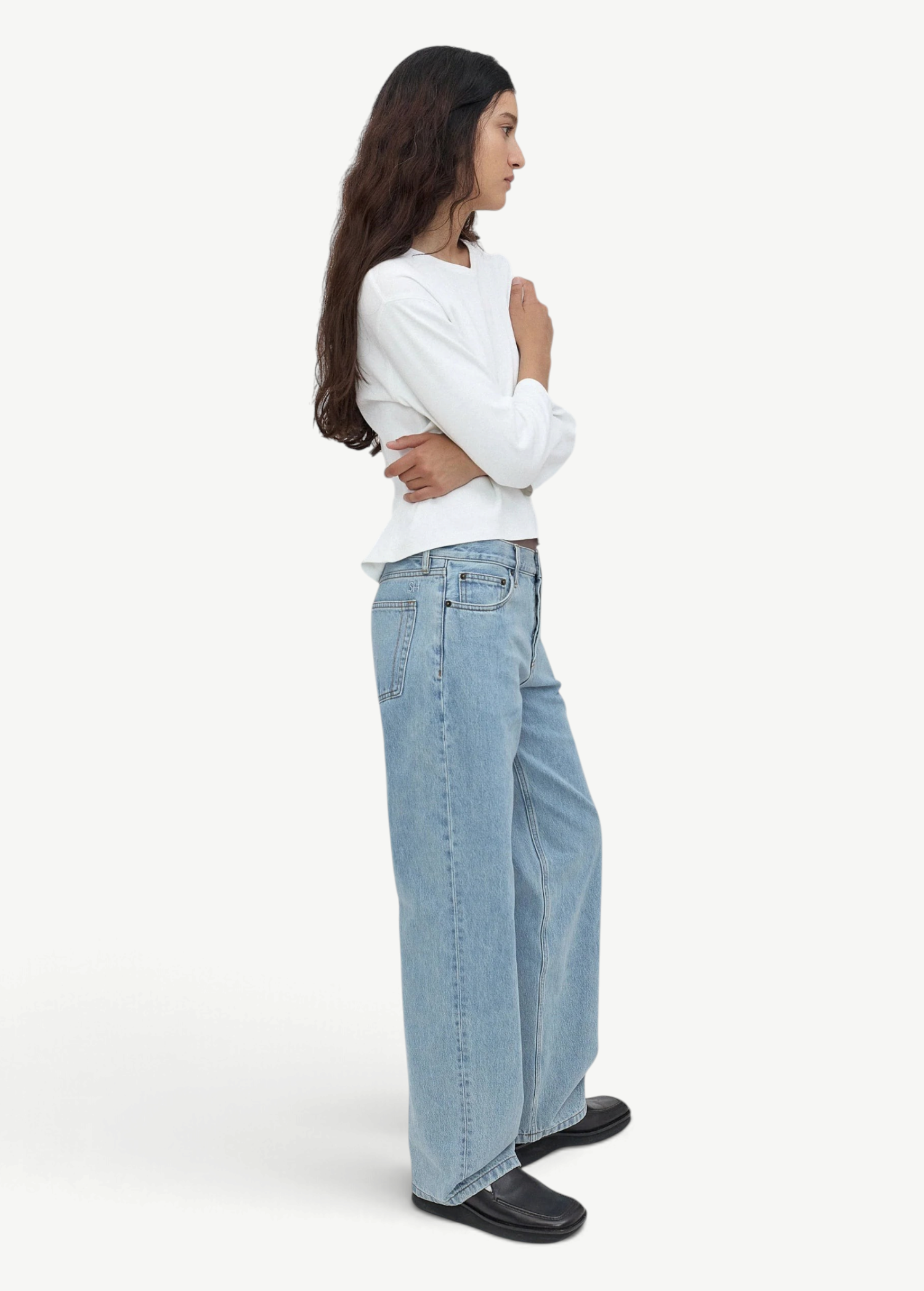 A woman with long dark hair stands in profile, wearing a white long-sleeve shirt, light blue Everyday Jeans featuring a relaxed silhouette, and black shoes against a plain light gray background.