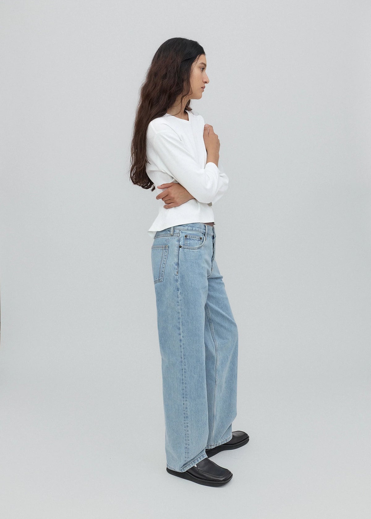 A woman with long dark hair stands in profile, wearing a white long-sleeve shirt, light blue Everyday Jeans featuring a relaxed silhouette, and black shoes against a plain light gray background.