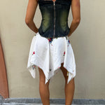 A person stands facing a beige wall, wearing a dark denim corset, the Celia Dragouni x KABE Embroidered Mini Skirt—a white upcycled mini skirt with floral prints and a handkerchief hem—and brown shoes. Their arms display visible pinkish tattoos.