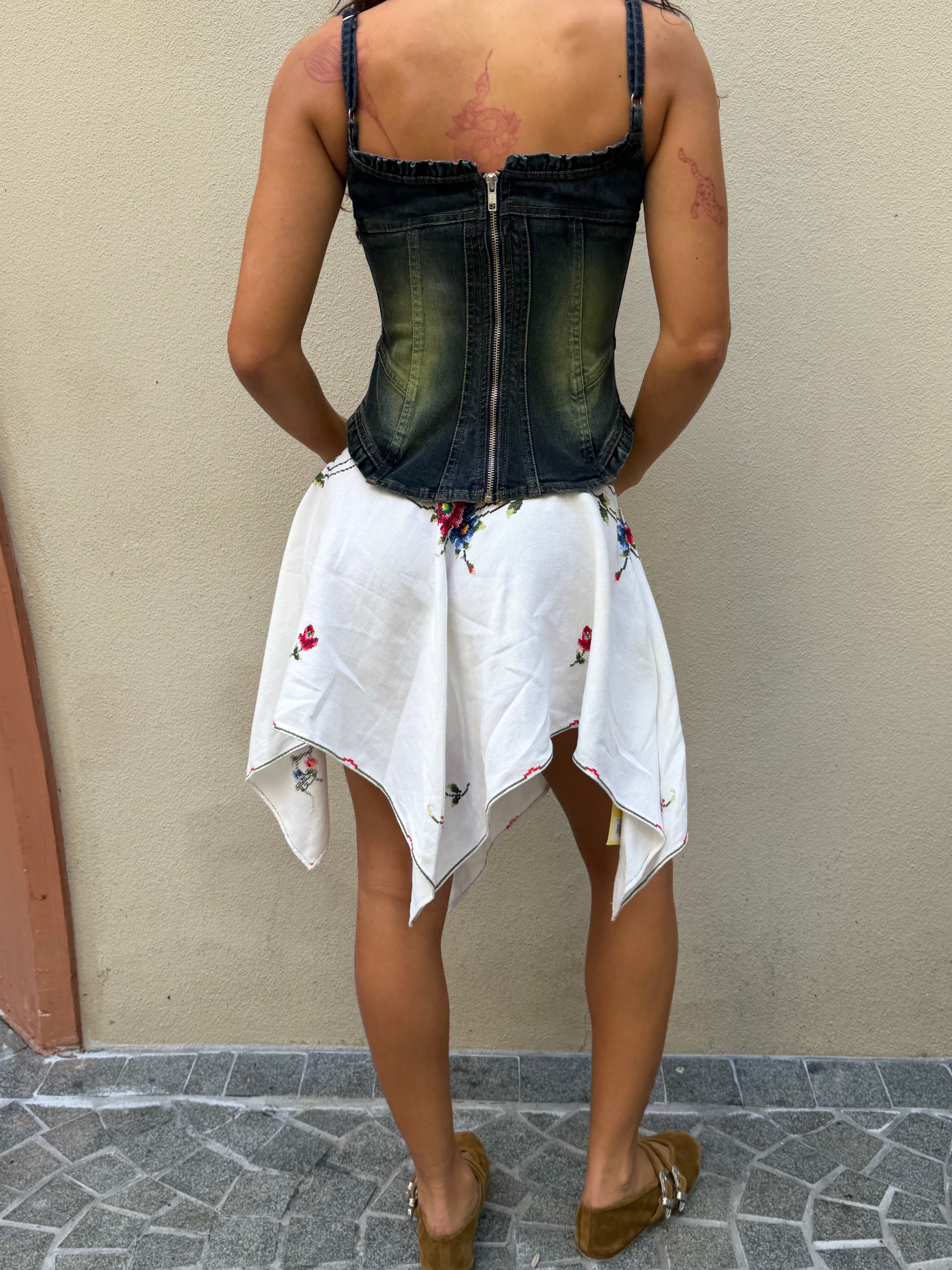 A person stands facing a beige wall, wearing a dark denim corset, the Celia Dragouni x KABE Embroidered Mini Skirt—a white upcycled mini skirt with floral prints and a handkerchief hem—and brown shoes. Their arms display visible pinkish tattoos.