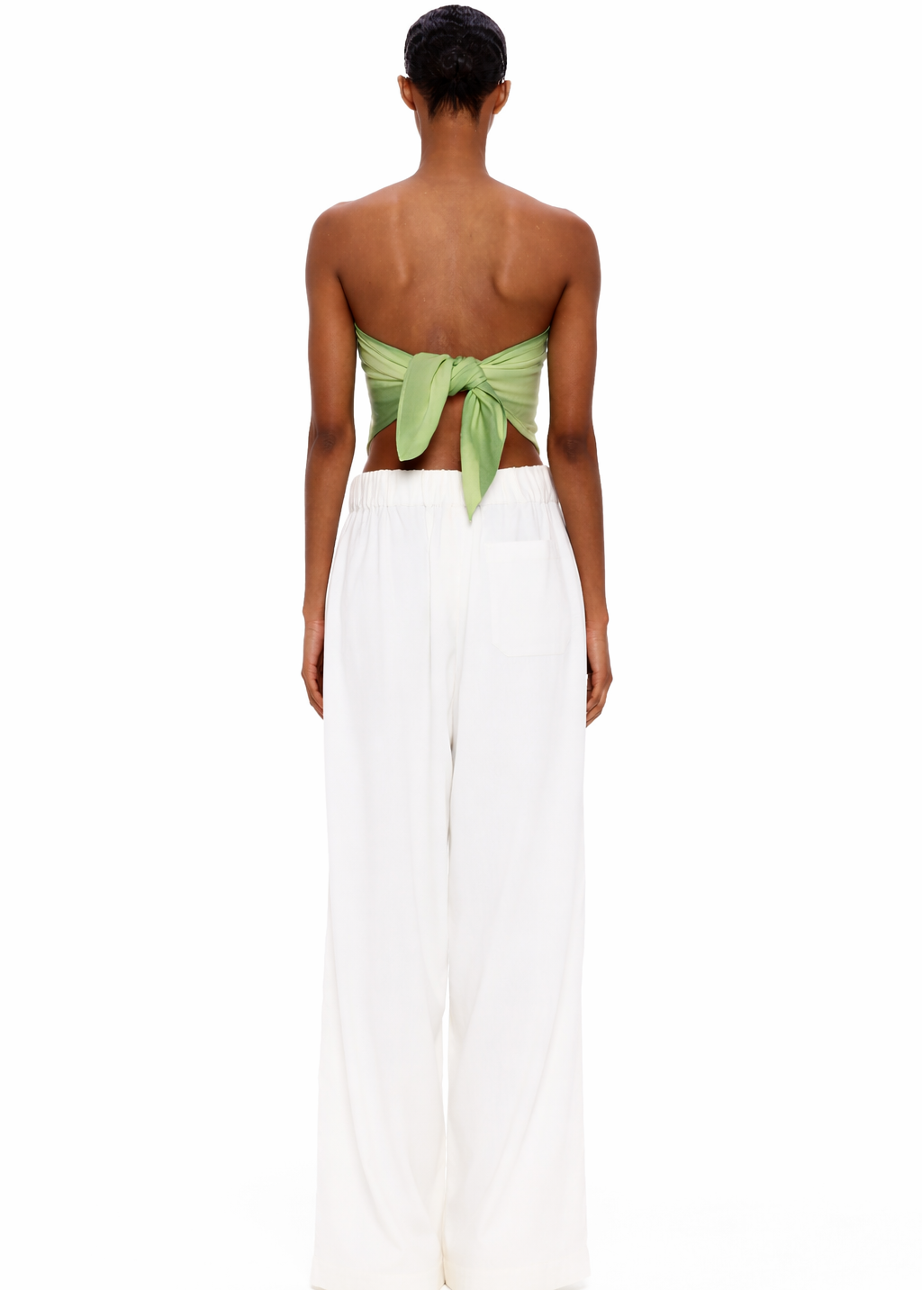 A person stands facing away, wearing The Triangle Top—a green back-tied bandeau inspired by traditional Moroccan weaving—and loose white pants, set against a plain white background.