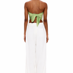 A person stands facing away, wearing The Triangle Top—a green back-tied bandeau inspired by traditional Moroccan weaving—and loose white pants, set against a plain white background.