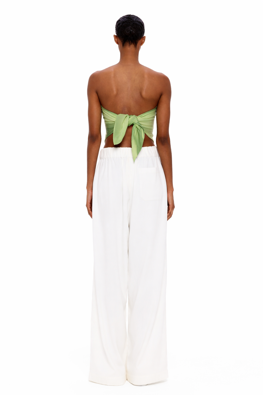 A person stands facing away, wearing The Triangle Top—a green back-tied bandeau inspired by traditional Moroccan weaving—and loose white pants, set against a plain white background.
