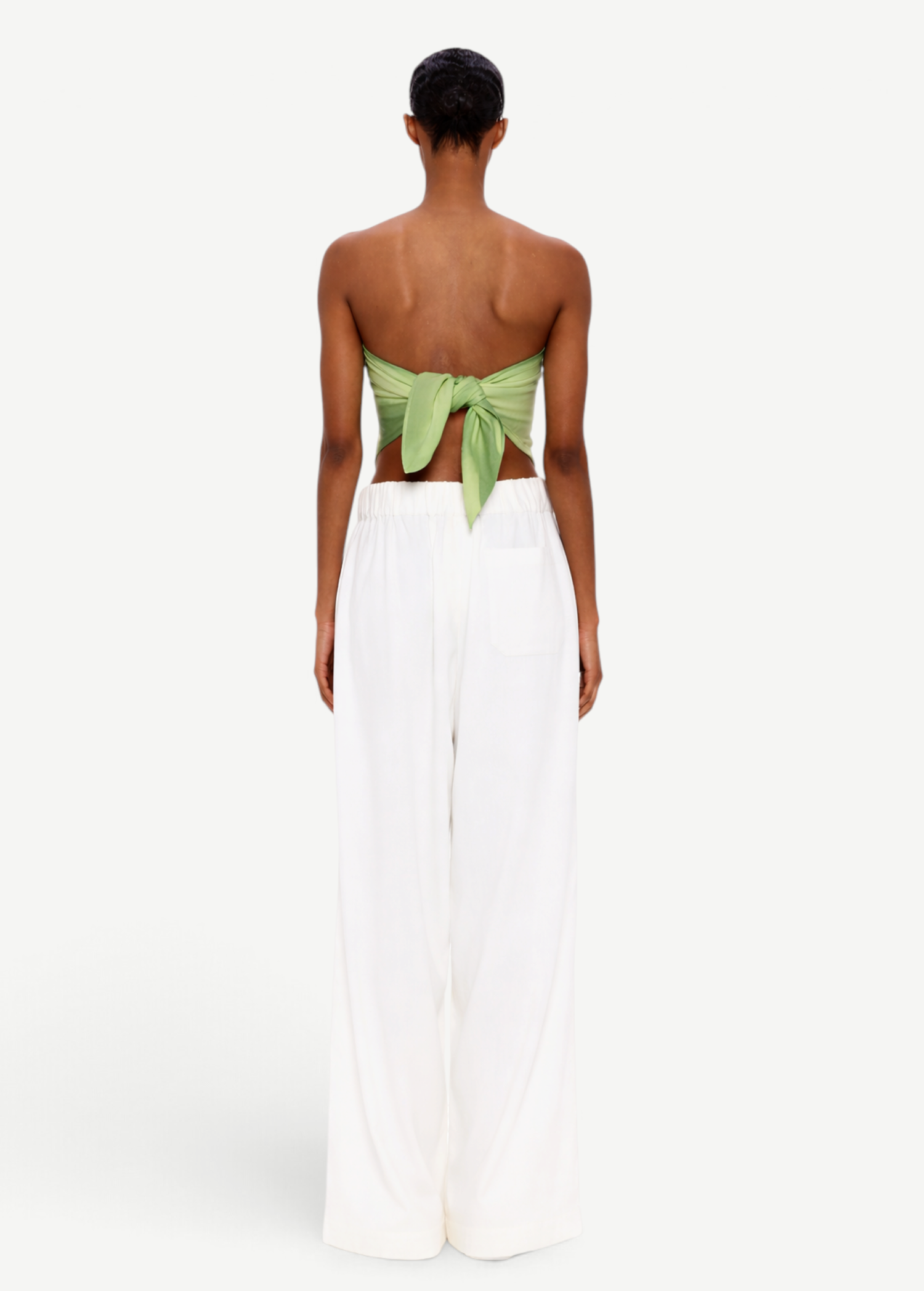 A person stands facing away, wearing The Triangle Top—a green back-tied bandeau inspired by traditional Moroccan weaving—and loose white pants, set against a plain white background.
