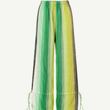The Parachute Pant features a wide-leg silhouette with vertical green, yellow, and white stripes, expertly crafted from handwoven cotton to highlight exquisite Moroccan craftsmanship. Designed with an elastic waistband and adjustable drawstrings at the ankles, these pants are showcased against a white background.