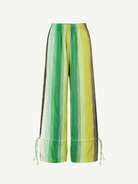 The Parachute Pant features a wide-leg silhouette with vertical green, yellow, and white stripes, expertly crafted from handwoven cotton to highlight exquisite Moroccan craftsmanship. Designed with an elastic waistband and adjustable drawstrings at the ankles, these pants are showcased against a white background.