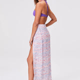 A woman stands sideways against a plain light background, looking forward. She wears a purple bikini top paired with the Sarong Skirt—a long, high-slit pastel skirt—along with flat sandals. Her long brown hair completes the effortlessly chic beachwear look.