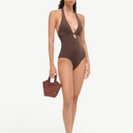 A woman wears a brown La Jolla Swimwear one-piece with a front cutout and metal ring detail, paired with black slide sandals and a small woven handbag for vacation dressing.