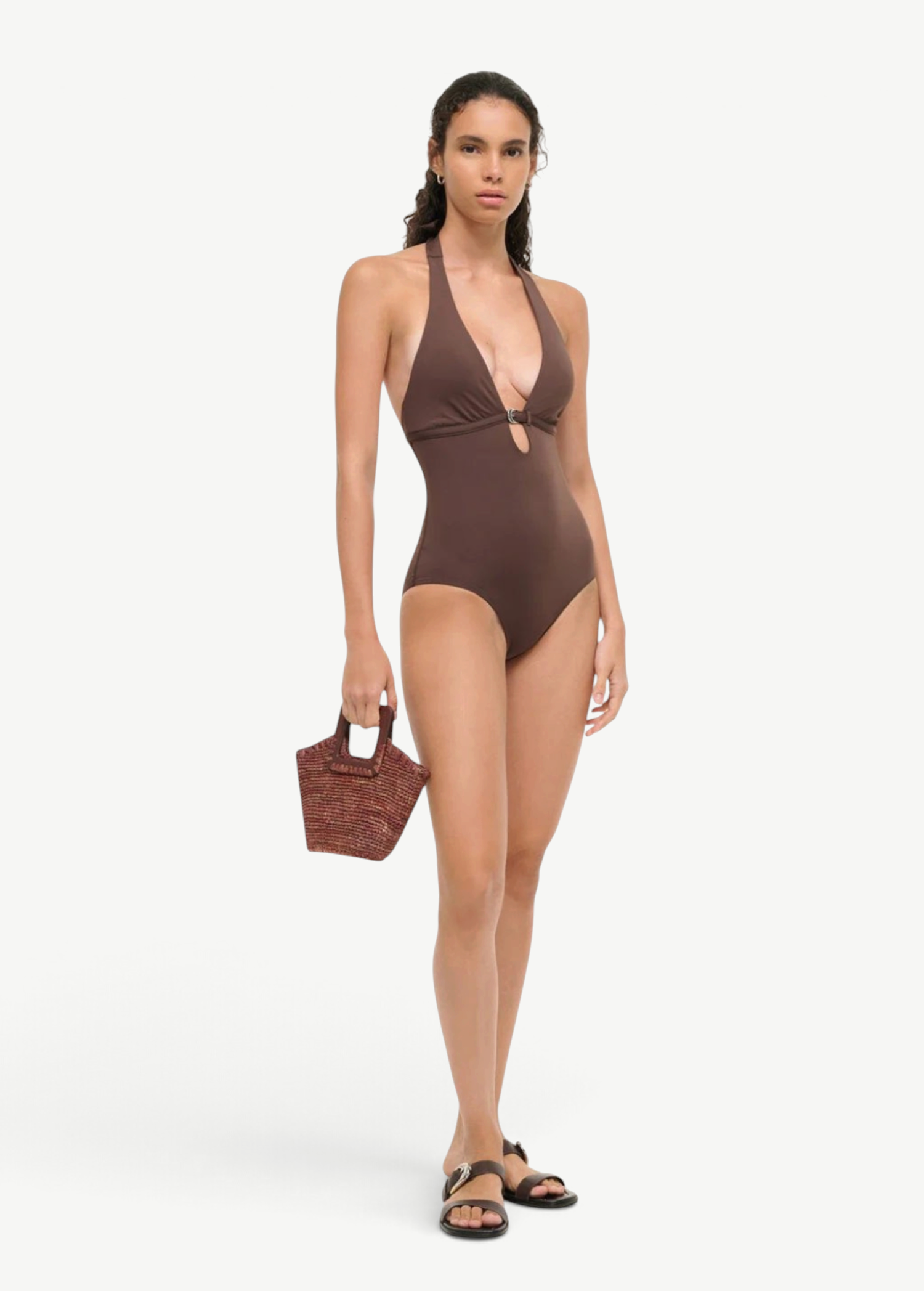 A woman wears a brown La Jolla Swimwear one-piece with a front cutout and metal ring detail, paired with black slide sandals and a small woven handbag for vacation dressing.