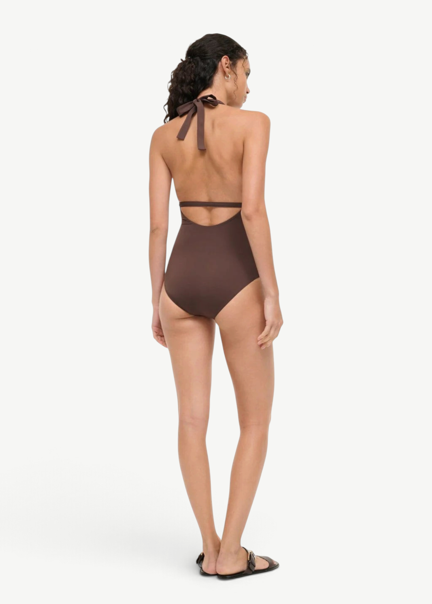 A woman viewed from behind wearing a brown La Jolla Swimwear one-piece swimsuit with a halter-style cut-out back and black flat sandals, perfect for vacation dressing.