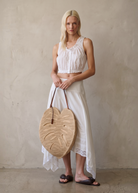 KABE SHOP INC large palm leaf shaped raffia bag with brown leather handles held by a blonde model in white summer wear