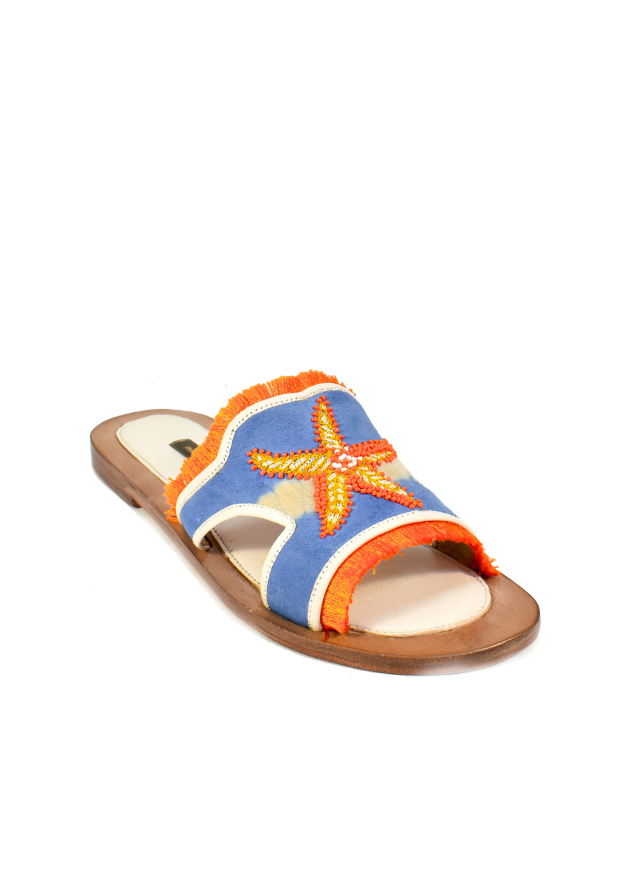 The Mazu Slide is a flat sandal featuring a blue upper with orange trim and a beaded starfish embellishment on top. Inspired by Silk Route artisan footwear, this open-toe sandal is finished with a tan sole, making it perfect for effortless summer style.