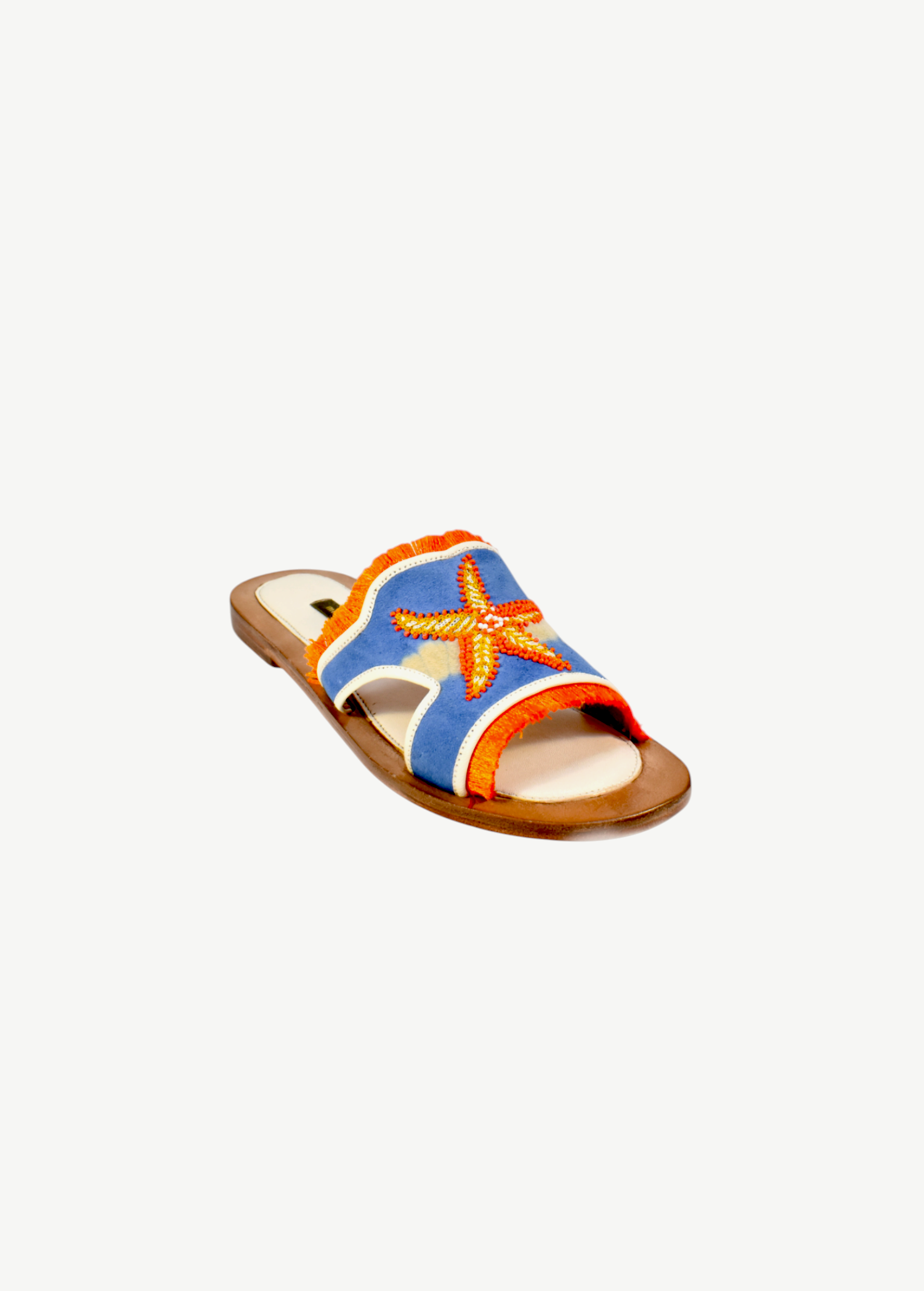 The Mazu Slide is a flat sandal featuring a blue upper with orange trim and a beaded starfish embellishment on top. Inspired by Silk Route artisan footwear, this open-toe sandal is finished with a tan sole, making it perfect for effortless summer style.