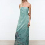 A model wears the Amara Dress, a strapless, light green midi with a sheer overlay featuring a printed coastal scene and scattered rhinestones.