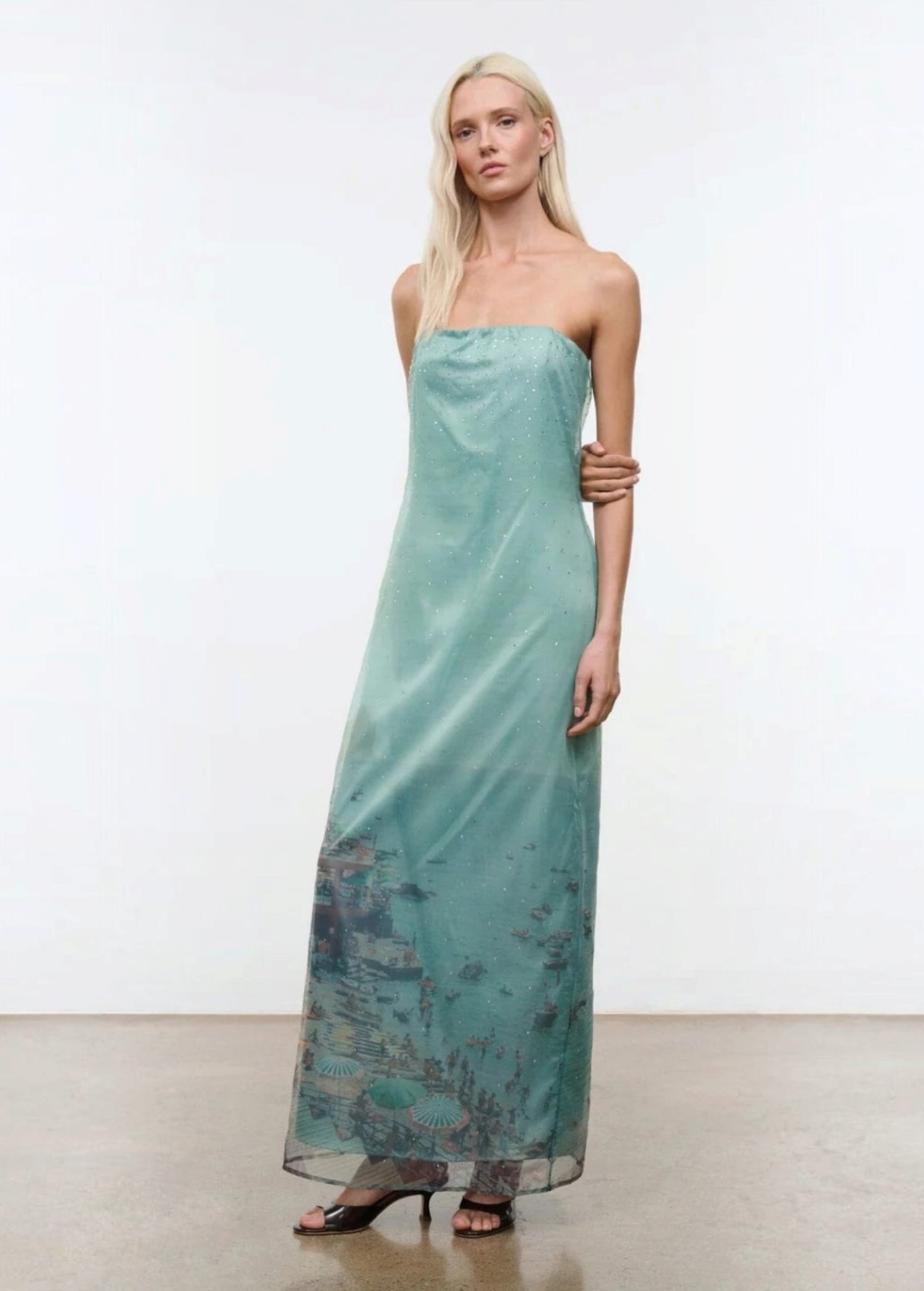 A model wears the Amara Dress, a strapless, light green midi with a sheer overlay featuring a printed coastal scene and scattered rhinestones.