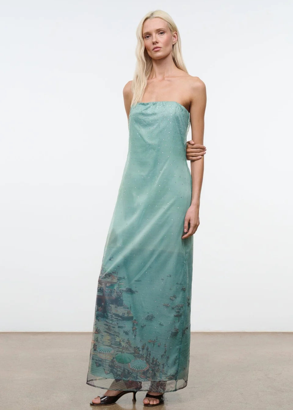 A model wears the Amara Dress, a strapless, light green midi with a sheer overlay featuring a printed coastal scene and scattered rhinestones.