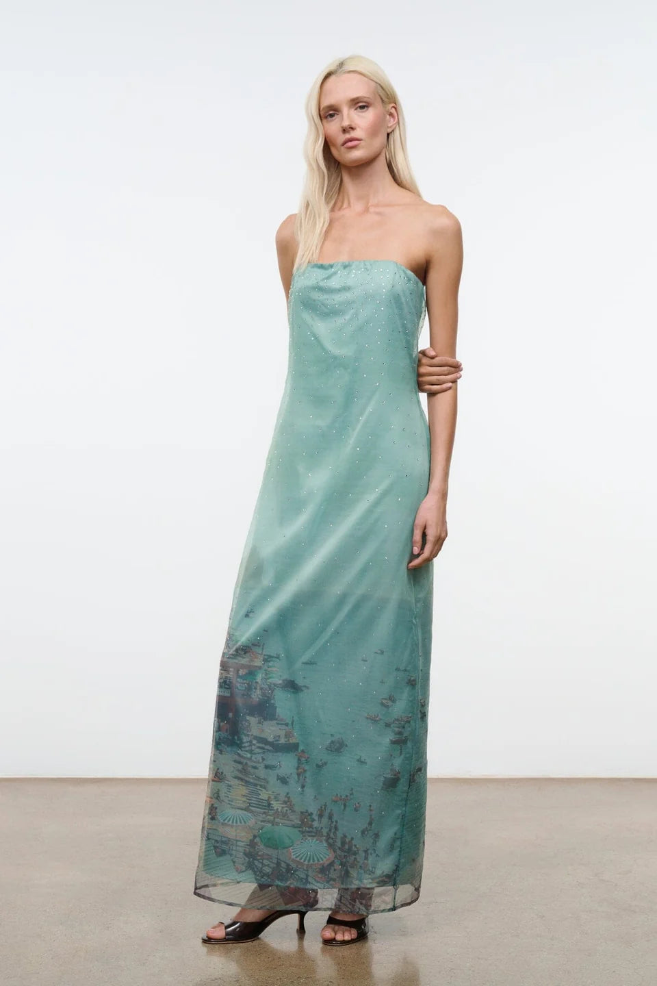 A model wears the Amara Dress, a strapless, light green midi with a sheer overlay featuring a printed coastal scene and scattered rhinestones.