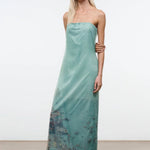 A model wears the Amara Dress, a strapless, light green midi with a sheer overlay featuring a printed coastal scene and scattered rhinestones.