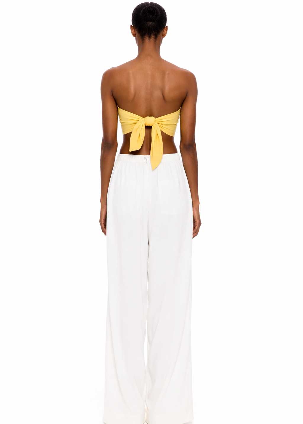 A person stands with their back to the camera, wearing The Triangle Top in yellow—a strapless design tied at the back—paired with loose white pants against a plain white background, showcasing a modern look inspired by sustainable Marrakech fashion.