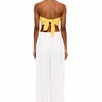 A person stands with their back to the camera, wearing The Triangle Top in yellow—a strapless design tied at the back—paired with loose white pants against a plain white background, showcasing a modern look inspired by sustainable Marrakech fashion.