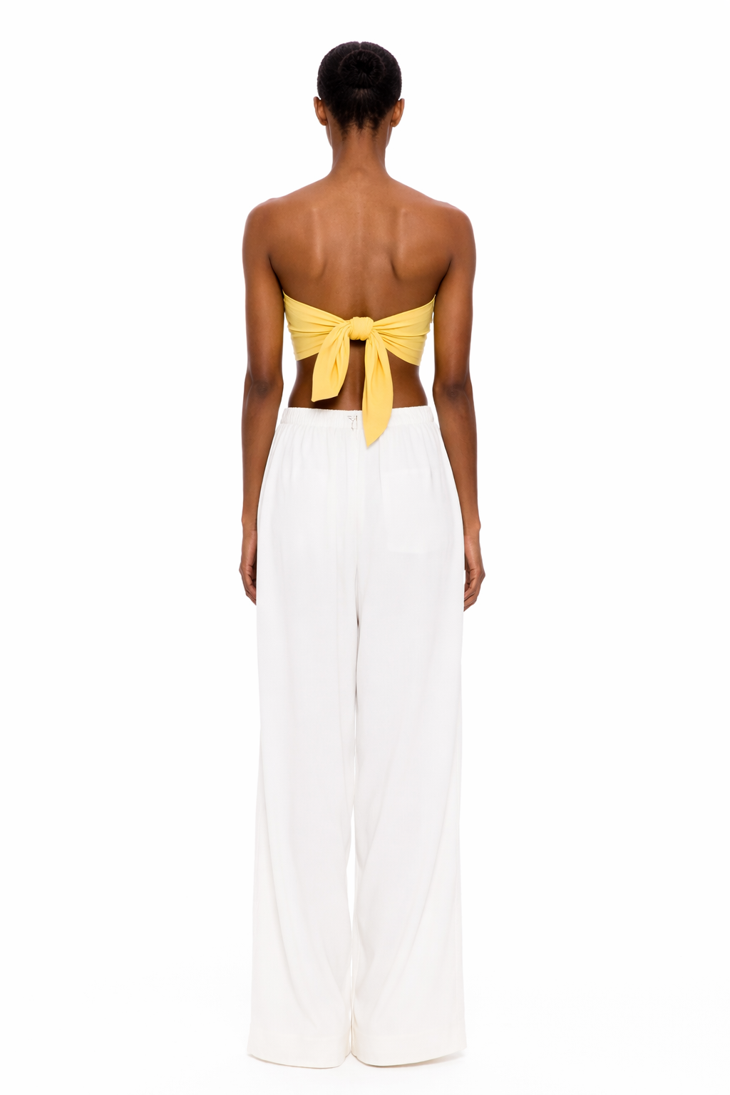 A person stands with their back to the camera, wearing The Triangle Top in yellow—a strapless design tied at the back—paired with loose white pants against a plain white background, showcasing a modern look inspired by sustainable Marrakech fashion.