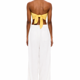 A person stands with their back to the camera, wearing The Triangle Top in yellow—a strapless design tied at the back—paired with loose white pants against a plain white background, showcasing a modern look inspired by sustainable Marrakech fashion.