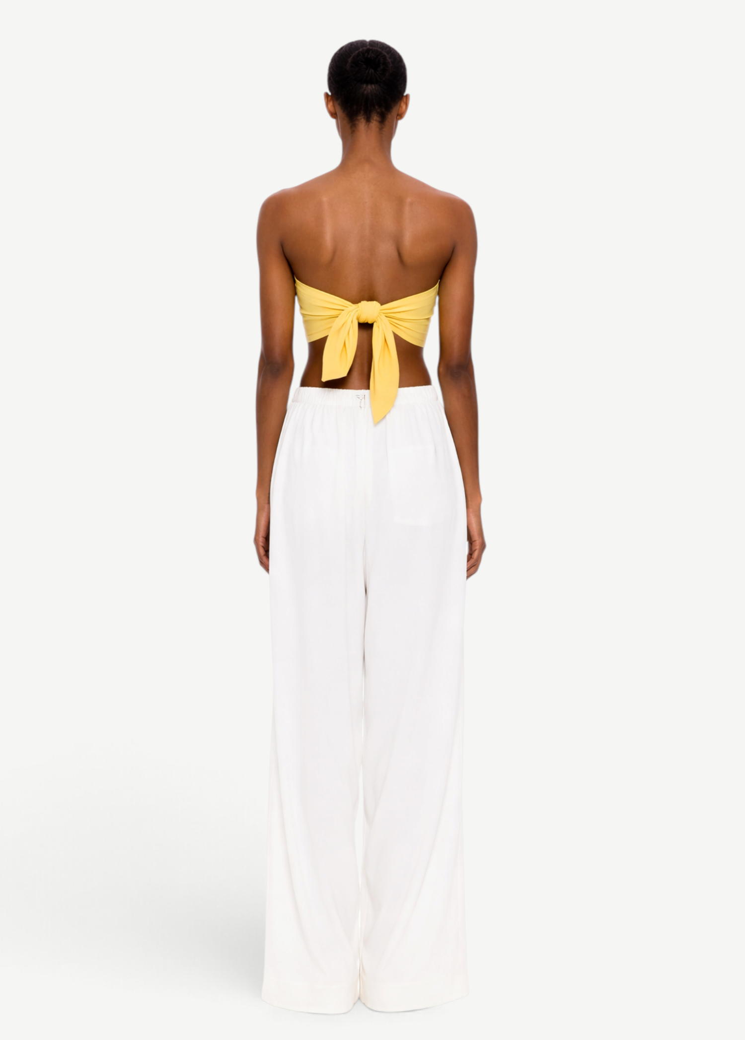 A person stands with their back to the camera, wearing The Triangle Top in yellow—a strapless design tied at the back—paired with loose white pants against a plain white background, showcasing a modern look inspired by sustainable Marrakech fashion.
