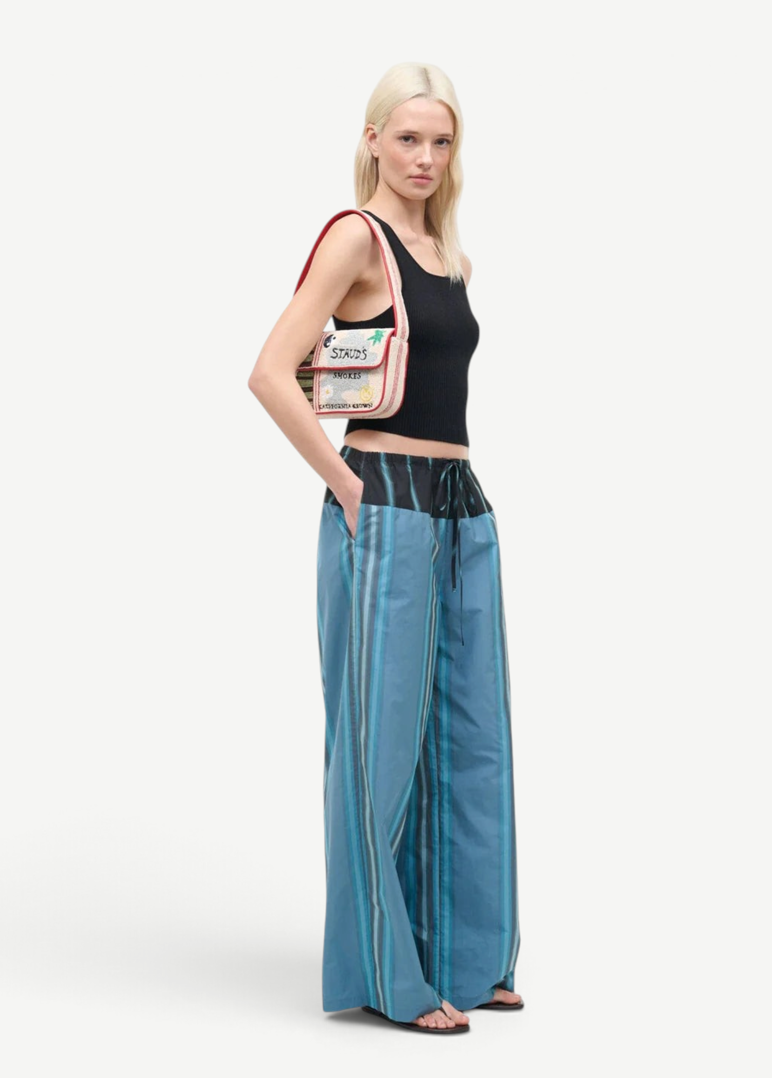 A person wearing a black tank top and Mateo Pant trousers with vertical blue stripes and a dark drawstring waistband, carrying a small, embroidered shoulder bag.