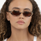 A person with curly hair is wearing the Sylvie sunglasses—an oval-shaped, tortoiseshell design featuring UV400 protection—and a white tank top, looking directly at the camera with a neutral expression.