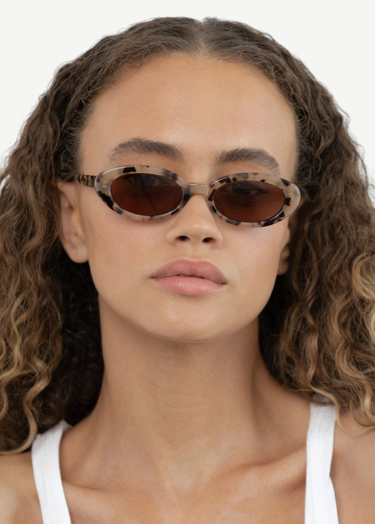 A person with curly hair is wearing the Sylvie sunglasses—an oval-shaped, tortoiseshell design featuring UV400 protection—and a white tank top, looking directly at the camera with a neutral expression.
