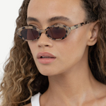 A woman with curly hair wears the Sylvie sunglasses, showcasing their patterned frame and UV400 protection, paired with a white tank top as she gazes slightly to the side against a plain background.