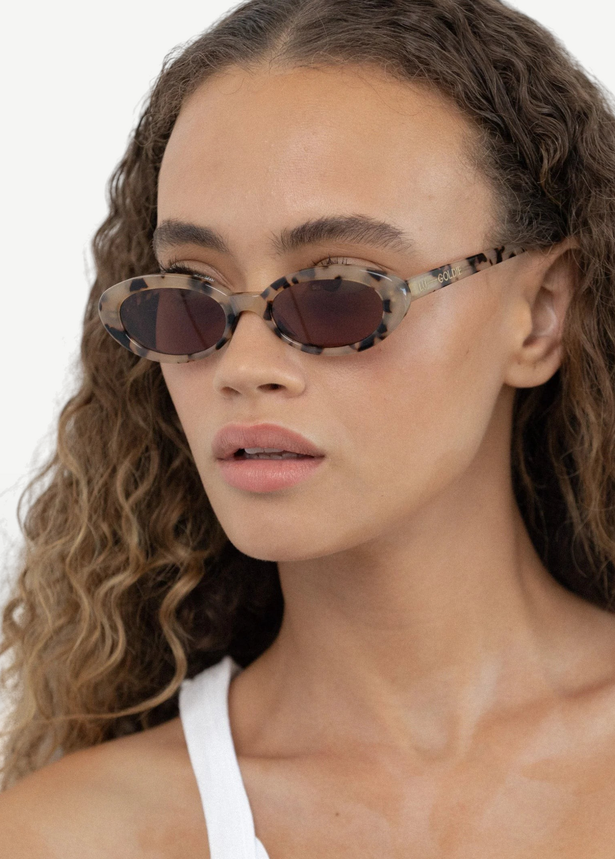 A woman with curly hair wears the Sylvie sunglasses, showcasing their patterned frame and UV400 protection, paired with a white tank top as she gazes slightly to the side against a plain background.