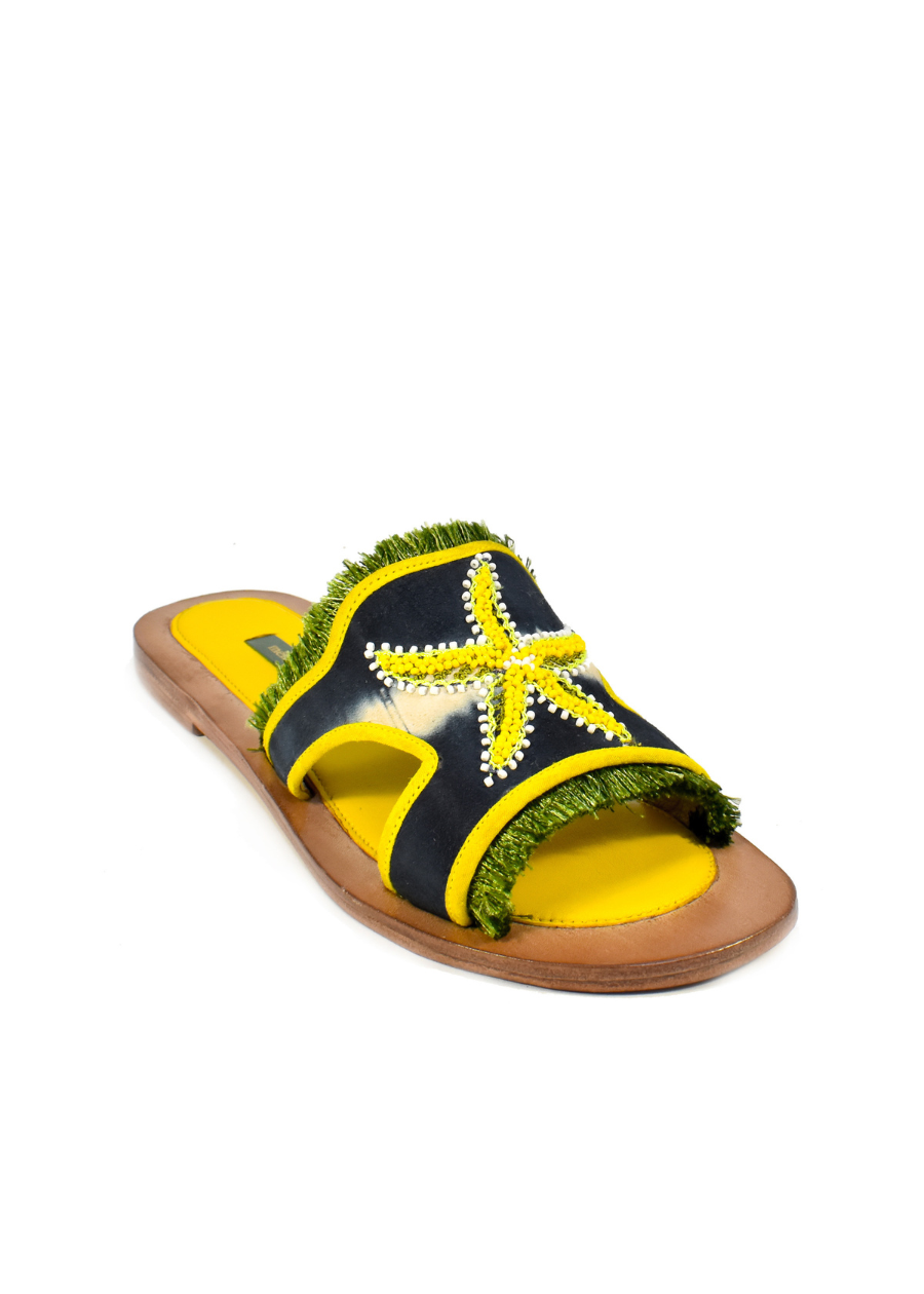 Introducing the Mazu Slide: these hand-embroidered leather sandals feature a brown sole, yellow footbed, and a black strap adorned with a yellow beaded starfish and green fringe accents—making them a stunning example of sustainable footwear.